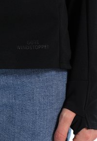 Manche noire Gore Windstopper avec trou pour le pouce sur le bras d'une personne appuyée contre un jean en denim bleu.