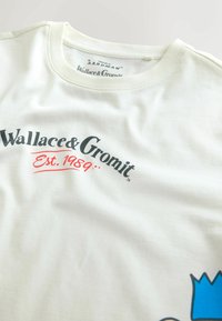 Tricou alb din bumbac cu un print grafic negru al "Wallace & Gromit" și "Est. 1989" în roșu, având un design cu o coroană albastră în partea de jos.