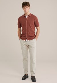 Camisa de manga corta de color óxido con un diseño acanalado texturizado, con botones en el frente, combinada con pantalones ligeros de color beige y mocasines negros.