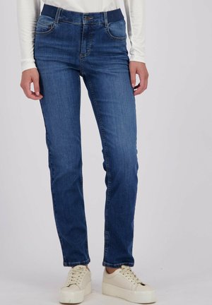 Vrouw in rechtvallende blauwe jeans, een witte lange mouwen top en witte platform sneakers tegen een effen achtergrond.