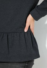 Top peplum di un grigio scuro realizzato in un tessuto morbido. Presenta un bordo arricciato e maniche aderenti. Mano posizionata lateralmente per la regolazione.