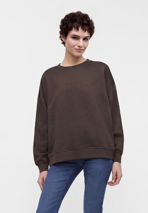 Jong persoon met kort krullend haar draagt een losse donkerbruine trui en blauwe jeans tegen een effen lichte achtergrond.