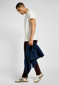 Camiseta blanca de manga corta, diseño ajustado, combinada con pantalones marrón oscuro. Sudadera azul marino caída sobre el brazo, zapatos beige con rayas negras y suelas de goma.