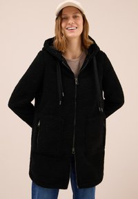 Femme souriante portant une casquette beige, un manteau polaire zippé noir avec de grandes poches avant, et un jean bleu devant un fond clair uni.