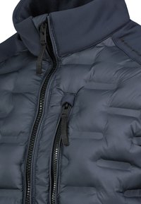 Dunkelblaue gepolsterte Jacke mit zwei schwarzen Reißverschlüssen, hohem Kragen und sichtbaren Nähten auf dem gesteppten Stoff.