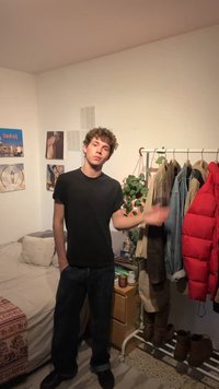 Jeune homme en t-shirt noir debout dans la chambre à côté d'un portant avec des manteaux et des bottes, un lit et un mur avec des photos.