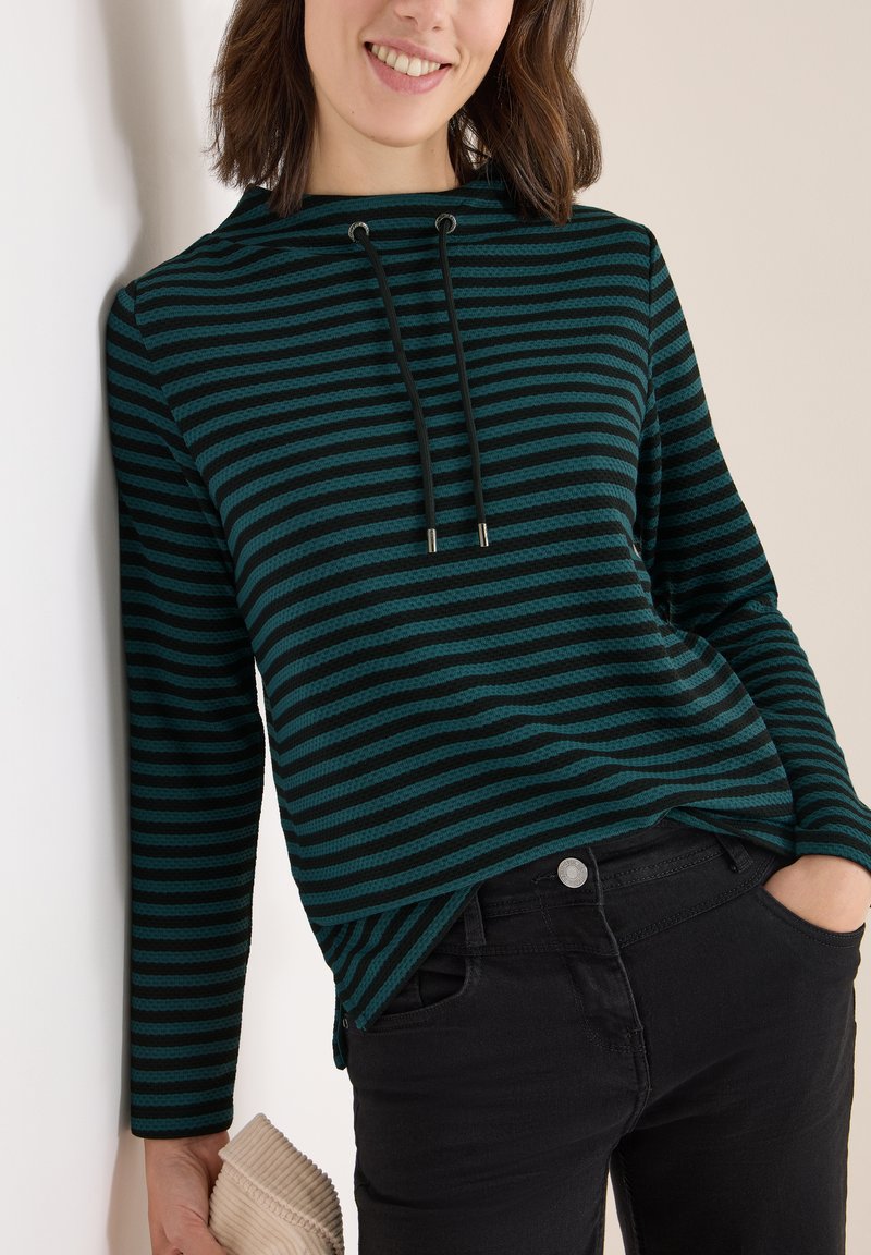 Pullover a righe in verde scuro e nero, con cappuccio, cordini neri e un motivo testurizzato, abbinato a jeans neri.