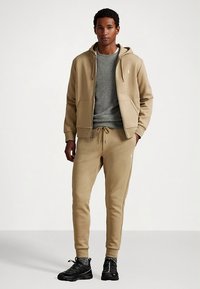 Beżowa bluza z kapturem z przednią kieszenią oraz pasujące joggery. Pod spodem strukturalny szary sweter i czarne buty sportowe.