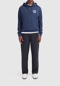 Marineblauwe hoodie met een grafisch detail aan de voorkant, gecombineerd met donkere broek met patchzakken en witte sneakers.