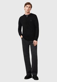 Sudadera con cremallera negra y cordones, con un bolsillo frontal. Combinada con pantalones de mezclilla oscuros y zapatos negros tipo slip-on. Diseño simple y casual.