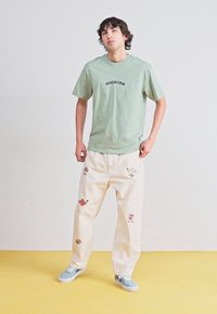Világos zöld pamut póló "DICKIES" felirattal, krémszínű nadrággal, amely színes hímzett motívumokkal díszített, és kék sportcipőkkel párosítva.