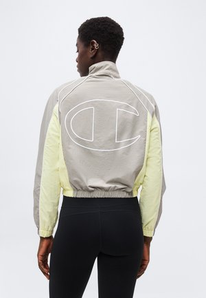 Persoană cu păr scurt și creț purtând o jachetă bej și galbenă scurtă, cu un logo mare conturat „C” pe spate și colanți negri.