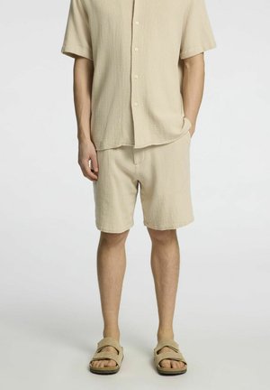 Beige strukturiertes Kurzarmhemd, kombiniert mit passender Shorts und hellen Slide-Sandalen, mit lockerer Passform und Knopfdetails.