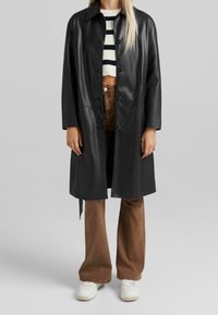 Czarny skórzany płaszcz o prostym kroju, z zapięciem na guziki i spiczastym kołnierzem. Połączony z pasiastym topem i brązowymi spodniami w stylu dzwonów.