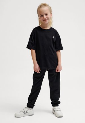 Svart oversized t-shirt med en liten vit logga på bröstet och svarta hoodiebyxor. Modellen bär vita sneakers med grå detaljer.