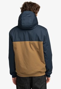 Hettejakke med fargeblokkdesign, mørkeblå skuldre og brun nedre del. Laget av et glatt, matt stoff.