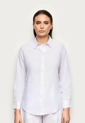 Donna con capelli castani lisci indossa una camicia bianca a maniche lunghe con bottoni, in piedi davanti a uno sfondo chiaro e uniforme.