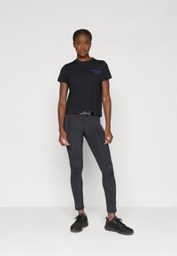 Juodas trumpas marškinėliai su mėlynu tekstu, derinami su priglundančiomis juodomis kelnaitėmis ir juodais sportiniais batais. Lygi medžiaga ir prisitaikanti forma.