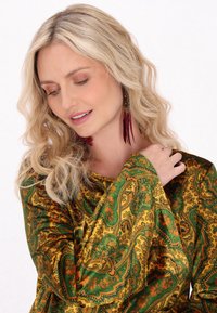 Top verde e oro con motivo paisley, maniche lunghe, caratterizzato da una texture liscia e un leggero lucido; adornato con lunghi orecchini bordeaux.