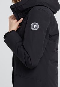 Sort polstret jakke med en tætsiddende hætte, ribbede manchetter og et rundt logo-lapel på venstre ærme. Glat, let materiale.