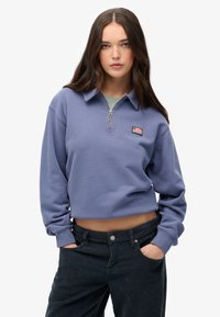 Sudadera corta de color lila con cremallera, cuello acanalado y parche de la marca. Combinada con pantalones de pana oscuros con bolsillos laterales.