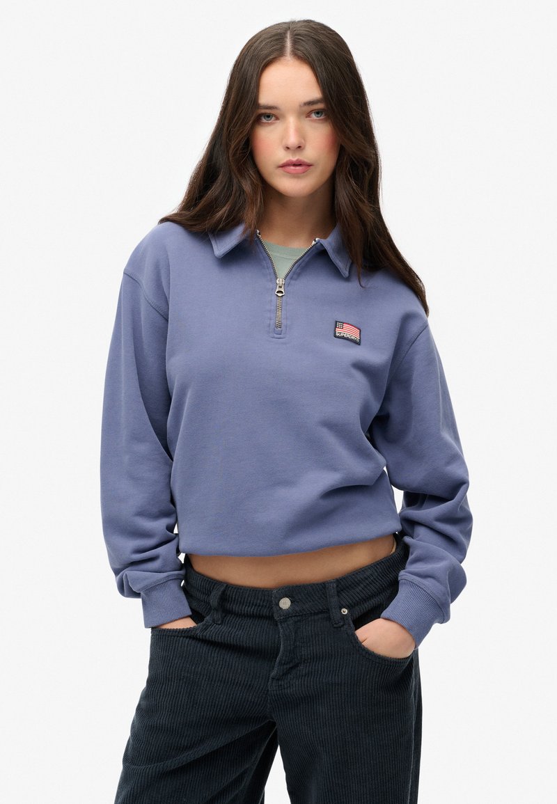 Sudadera corta de color lila con cremallera, cuello acanalado y parche de la marca. Combinada con pantalones de pana oscuros con bolsillos laterales.