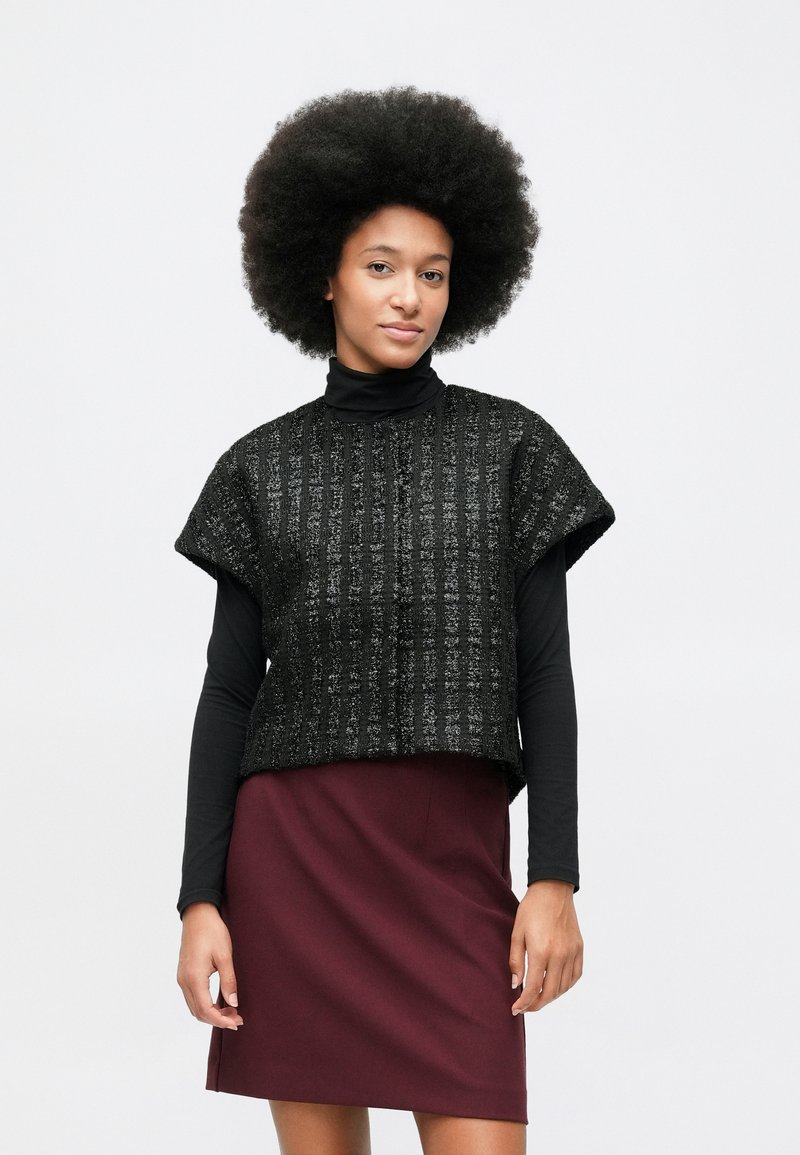 Žena s afro frizurom nosi crni kratki pleteni pulover s kratkim rukavima preko crne bluze s dugim rukavima i bordo suknju.