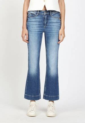 Frau trägt mittelhohe, hellblaue, ausgestellte Jeans mit hochgekrempelten Säumen und weißen Turnschuhen, steht vor einem schlichten weißen Hintergrund.