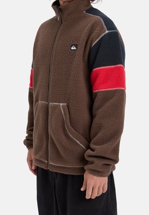 Mann trägt braune Fleecejacke mit schwarz-roten Farbakzenten an den Ärmeln, Frontreißverschluss und zwei Fronttaschen, steht vor weißem Hintergrund.