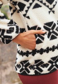 Fleece pullover med sort og hvidt geometrisk mønster, der har en lomme på siden og et blødt, tekstureret stof.