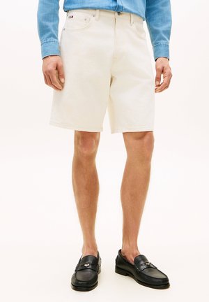 Mand iført off-white knælange shorts, en lyseblå denimskjorte og sorte læderloafers, stående foran en ensfarvet baggrund.