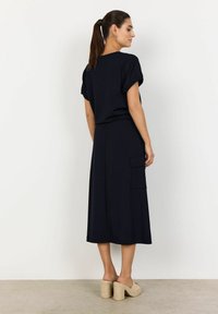 Navyblaues Kleid mit kurzen, umgeschlagenen Ärmeln, in mittellanger Knielänge und zwei seitlichen Taschen. Glatter Stoff mit einer lockeren Passform. Chunky-Absatzsandalen.