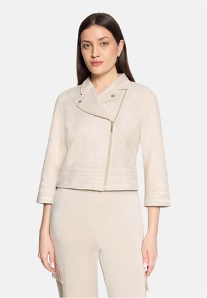 Vrouw draagt een licht beige cropped jack met een asymmetrische rits aan de voorkant en bijpassende beige broek tegen een effen achtergrond.