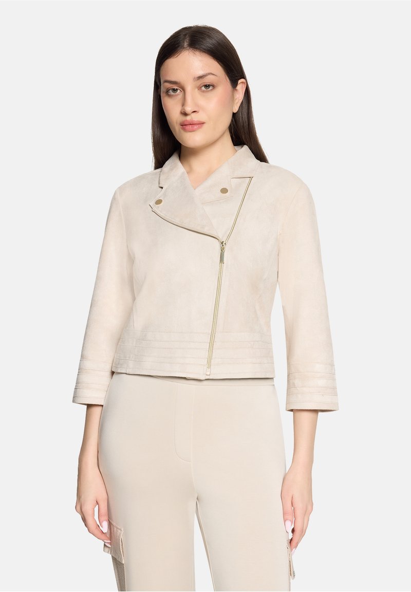 Femme portant une veste courte beige clair avec une fermeture éclair asymétrique à l'avant et un pantalon beige assorti sur un fond uni.