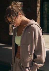 Köisiga kapuutsiga tõmblukuga sweatshirt helesinises toonis, lahtise lõikega, ees taskud ja valge logo. Kandmisel on see helesinise lühikeste käistega T-särgi peal, sobitatuna ühtivate pükstega.