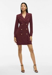 Žena v burgundském dvouřadém saku šatů se zlatými knoflíky a černými high-heeled sandály, stojící na neutrálním pozadí.