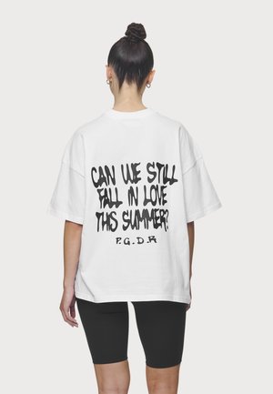 Kobieta z włosami upiętymi w kok, ubrana w białą oversize koszulkę z czarnym napisem „Czy nadal możemy się zakochać tego lata? P.G.D.R” na plecach oraz czarne szorty.
