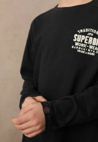 Cămașă neagră cu mânecă lungă, cu guler rotund, detalii cu logo alb pe piept și etichetă pe manșetă. Material din bumbac, textură moale.