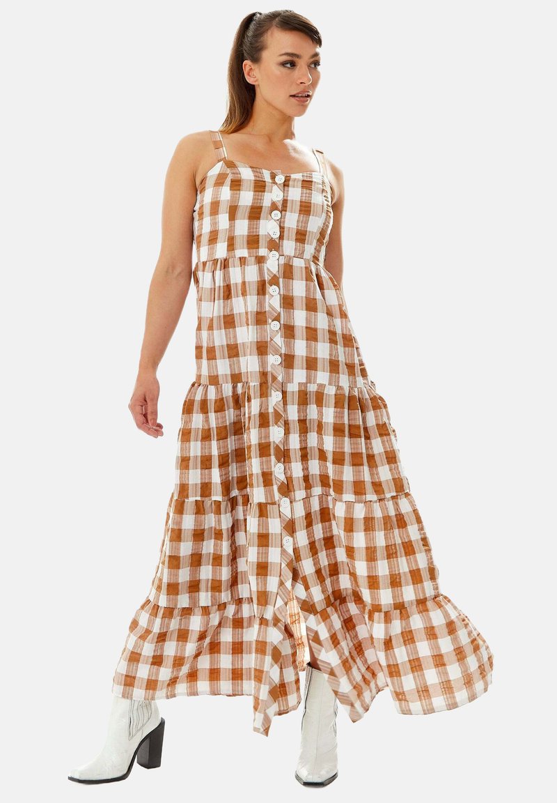 Maxi dress in brown and white checkered pattern, with a square neckline, tiered design, and front button closure. Worn with white boots.
