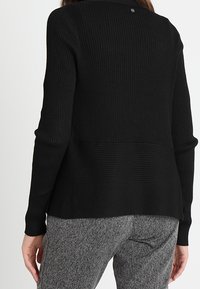 Czarny prążkowany sweter z wysokim kołnierzem i teksturowanym wzorem, z obniżonym dołem. Z tyłu znajduje się małe logo w pobliżu krawędzi szyi.