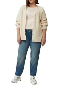 Creme gebreide cardigan over een beige en witte gestreepte t-shirt, gecombineerd met blauwe rechte jeans en lichtgrijze sneakers.