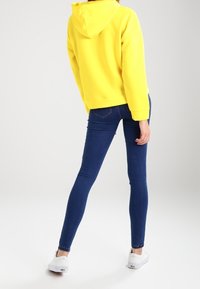 Felpa gialla con cappuccio e tasca a marsupio, abbinata a jeans skinny blu scuro e sneakers bianche. Texture liscia e design casual.
