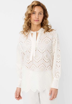 Blusa bianca leggera con ricamo ad occhio, scollo rotondo, maniche lunghe e dettaglio con fiocco frontale. Orlo ondulato.