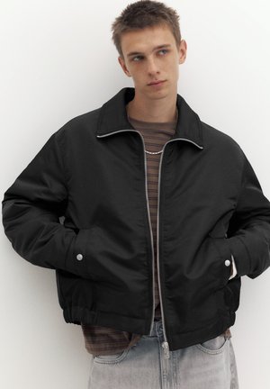 Jeune homme portant une veste noire zippée sur une chemise rayée, debout les mains dans les poches devant un fond blanc uni.