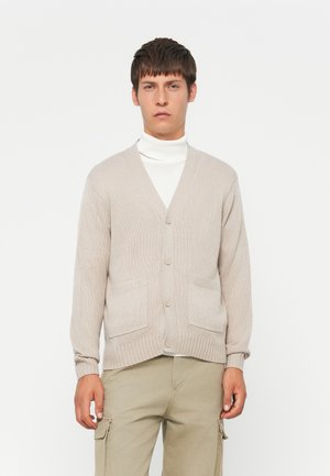 Young man wearing beige buttoned cardigan over white turtleneck and beige cargo pants, standing against plain white background.