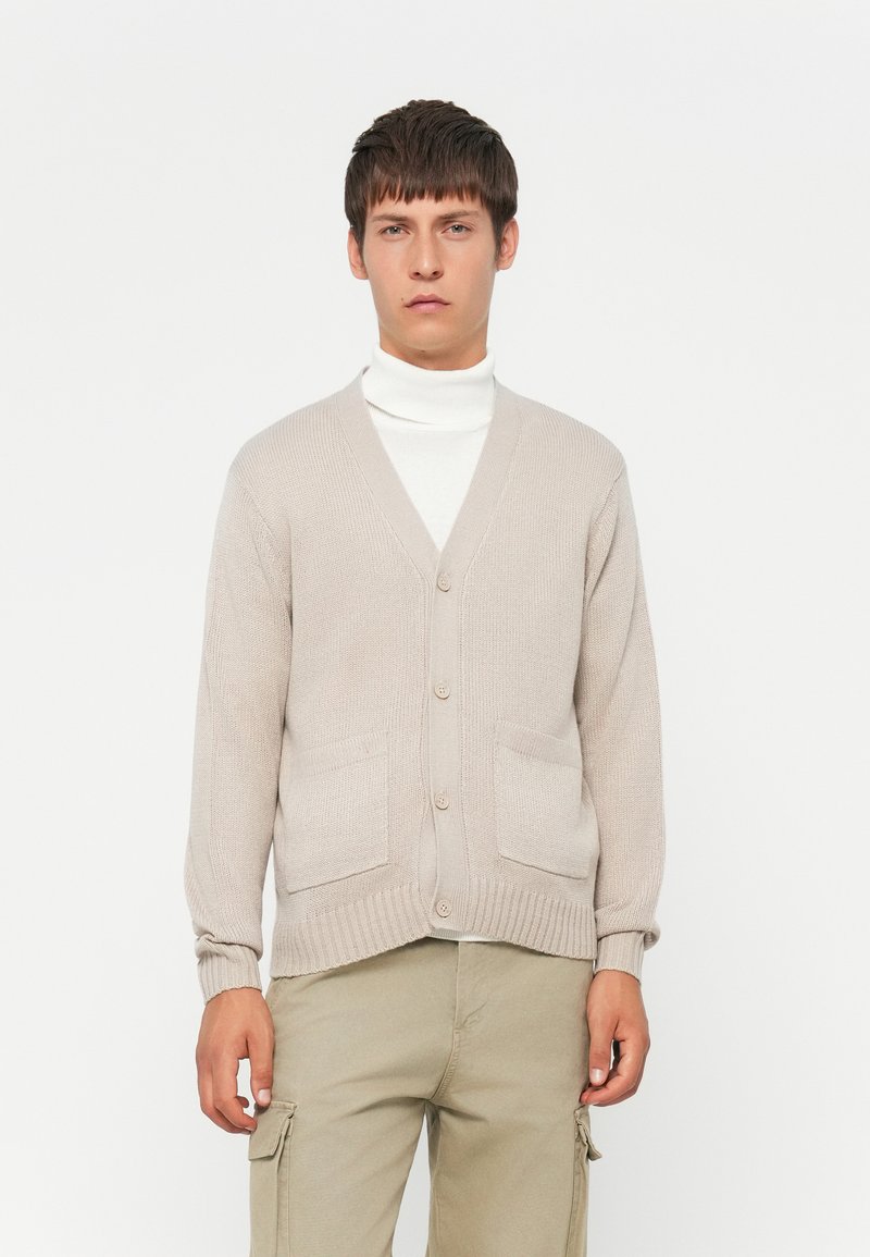 Ungdommelig mann med beige knappet cardigan over hvit turtleneck og beige cargo-bukser, stående mot en ensfarget hvit bakgrunn.