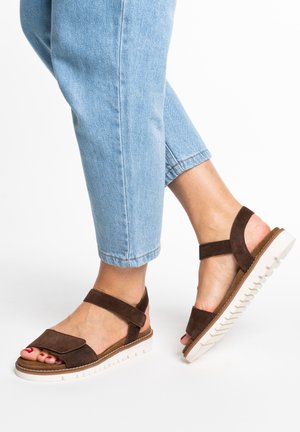 Person wearing light blue jeans and brown suede sandals with white ridged soles, one foot slightly raised against white background.