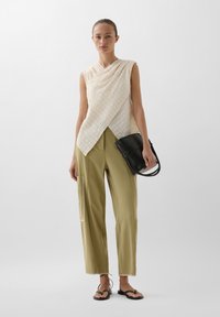 Cremfarbener ärmelloser Top mit asymmetrischem Fall, kombiniert mit olivgrünen Hosen mit seitlichen Taschen und dekorativen Nähten. Schwarze Handtasche.