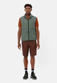 Gilet verde senza maniche con zip frontale e tasche, indossato sopra una camicia arancione a maniche corte, abbinato a pantaloni cargo marroni e scarpe nere.