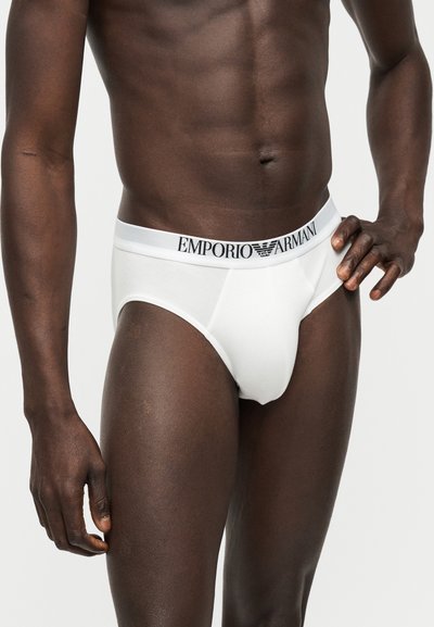 Ανδρικός κορμός φορώντας λευκά εσώρουχα Emporio Armani, στέκεται με το ένα χέρι στη μέση μπροστά σε μονόχρωμο φόντο.